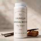 A white plastic bottle of DST Fragrances Vanilla Sandalwood Dry Shampoo, styled on a neutral surface with vanilla beans and a wood cube in the background.