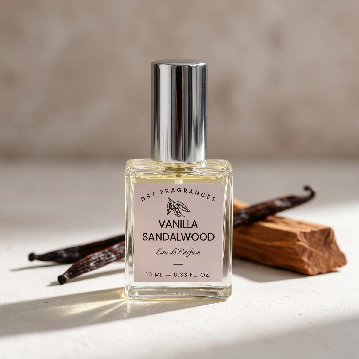 A 10 ml bottle of DST Fragrances Vanilla Sandalwood Eau de Parfum, featuring a clear glass bottle with a silver cap and a light pink label, styled with vanilla beans and a piece of wood on a neutral beige background.