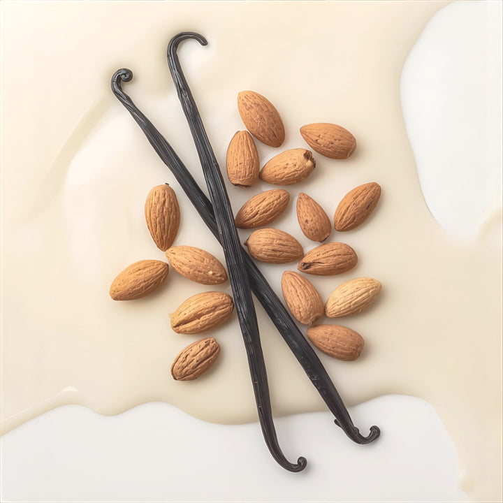 Two dark vanilla bean pods and several whole almonds arranged on a creamy off-white liquid background.