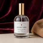 A clear glass bottle of DST Fragrances Velvet Ash Room & Linen Spray with a white label and a metallic gold cap, set against a dark burgundy velvet background with a beige textured fabric prop.