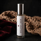A 10 ml roll-on bottle of 'Velvet Ash' diffuser oil by DST Fragrances, featuring a white label and a reflective silver cap, set against a dark background with a brown crochet blanket and a red velvet cloth.
