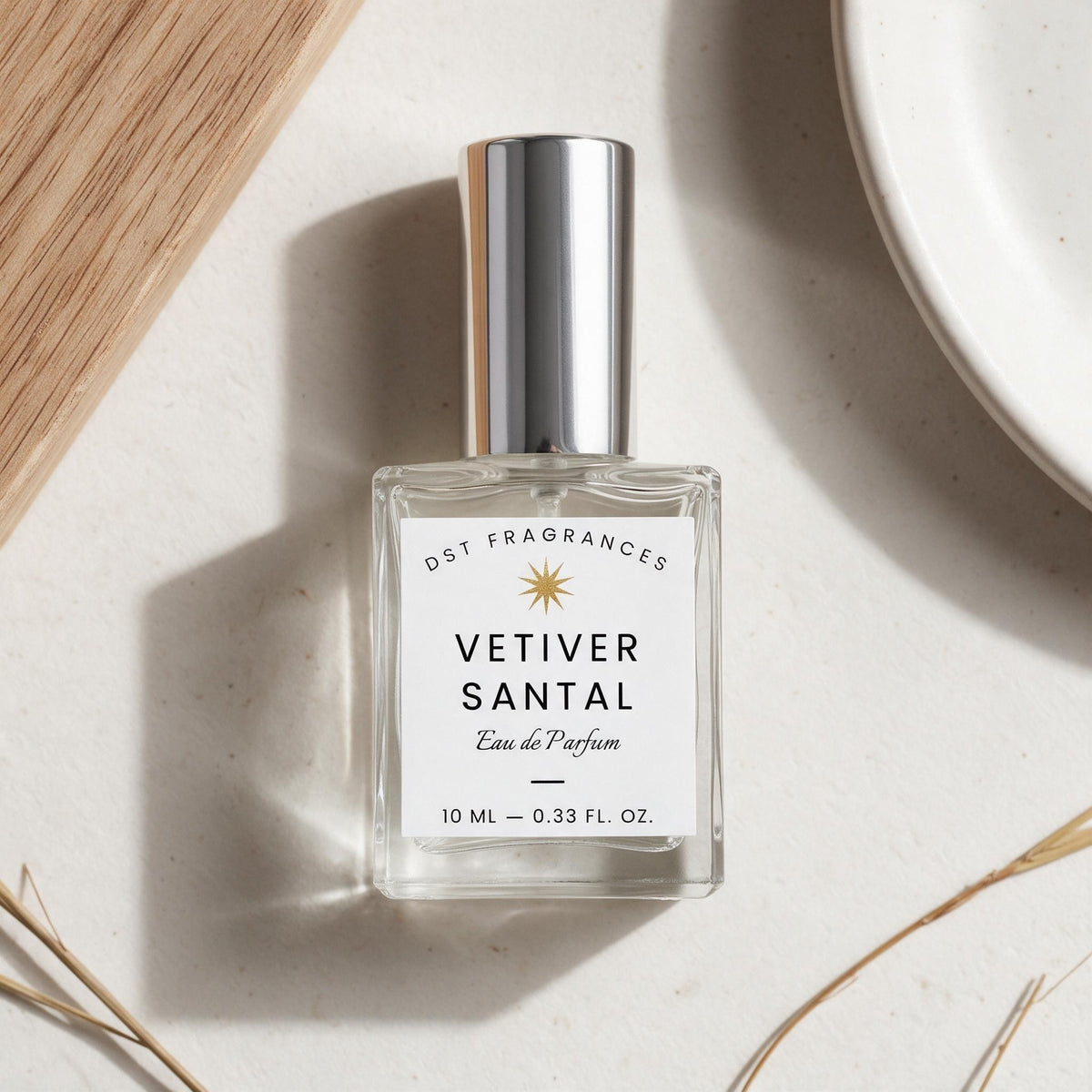 A 10 ml bottle of DST Fragrances Vetiver Santal Eau de Parfum, featuring a clear glass body, a white label with a gold star logo, and a reflective silver cap, styled on a textured beige surface with a wooden slice and a white plate.