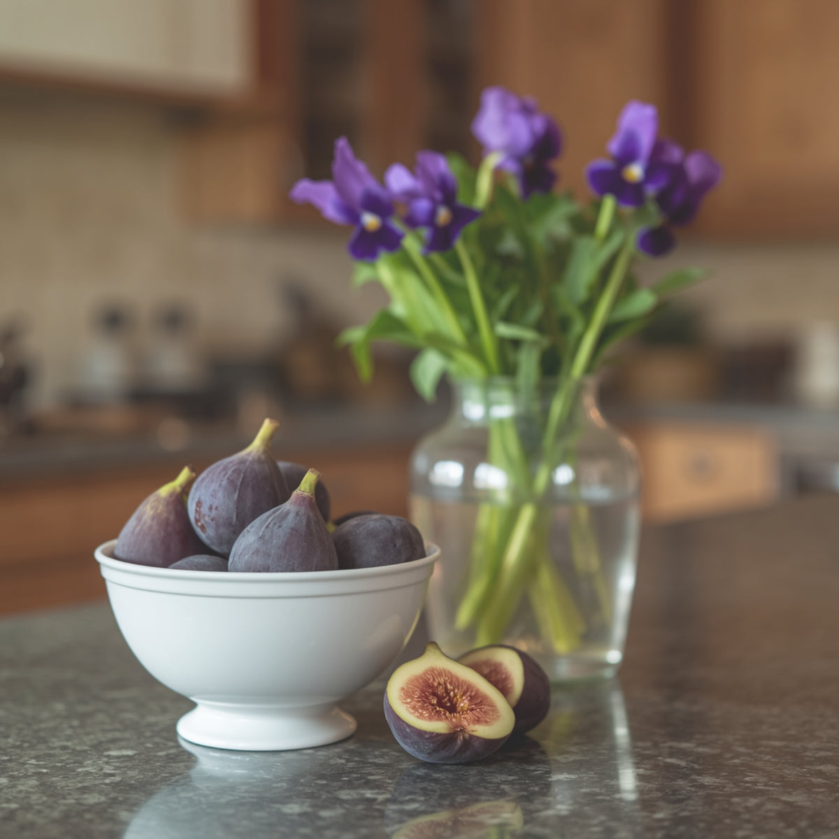 A white ceramic bowl filled with whole and halved figs sits on a kitchen countertop, with a glass vase of purple flowers blurred in the background.