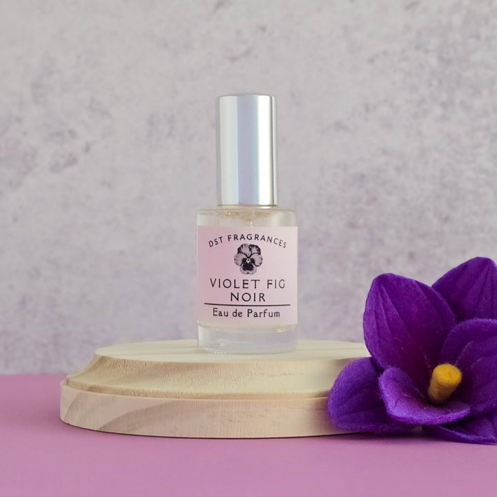 Perfume bottle labeled 'Violet Fig Noir' on a wooden stand with a purple flower against a light gray background.