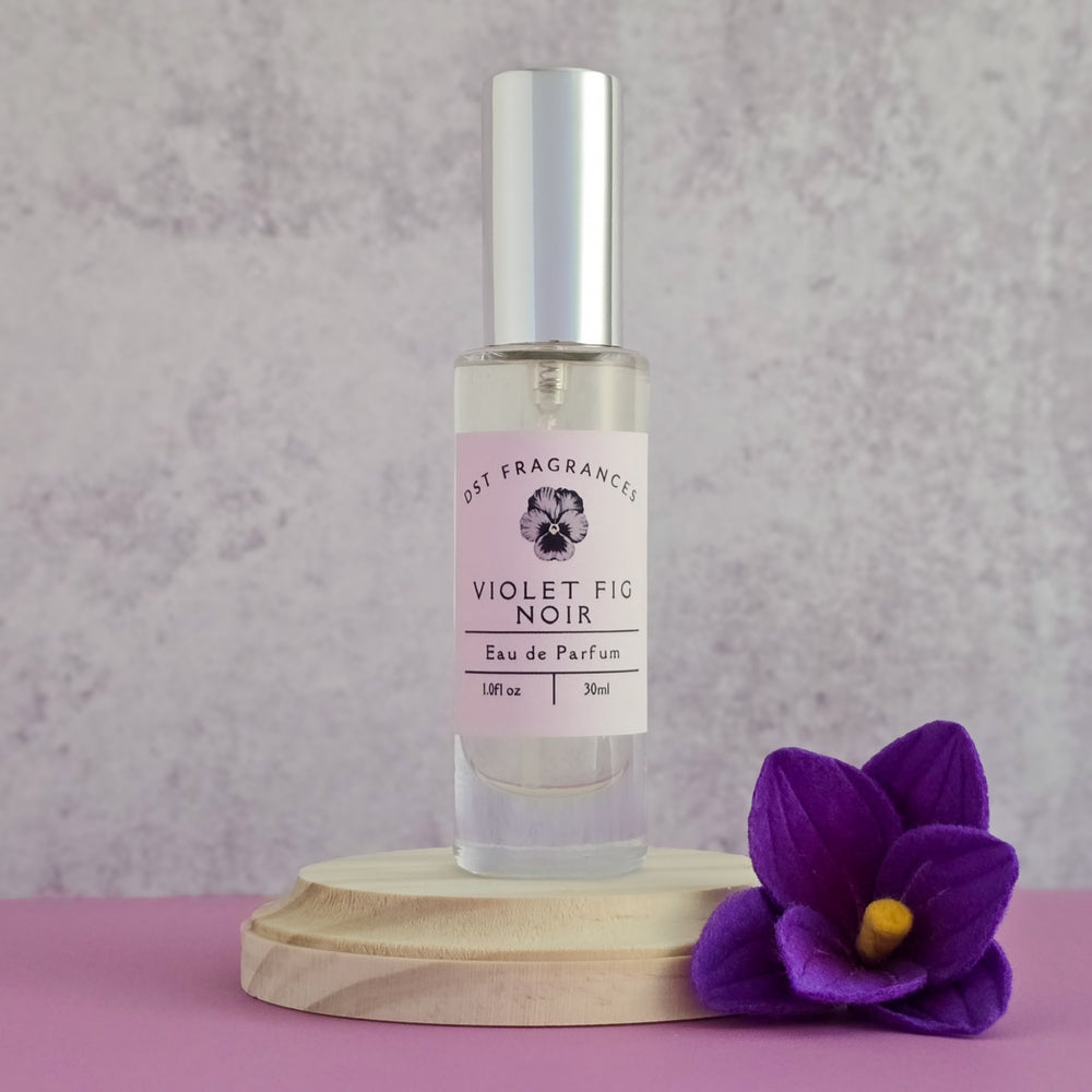 Perfume bottle labeled 'Violet Fig Noir' on a wooden stand with a purple flower against a light gray background.