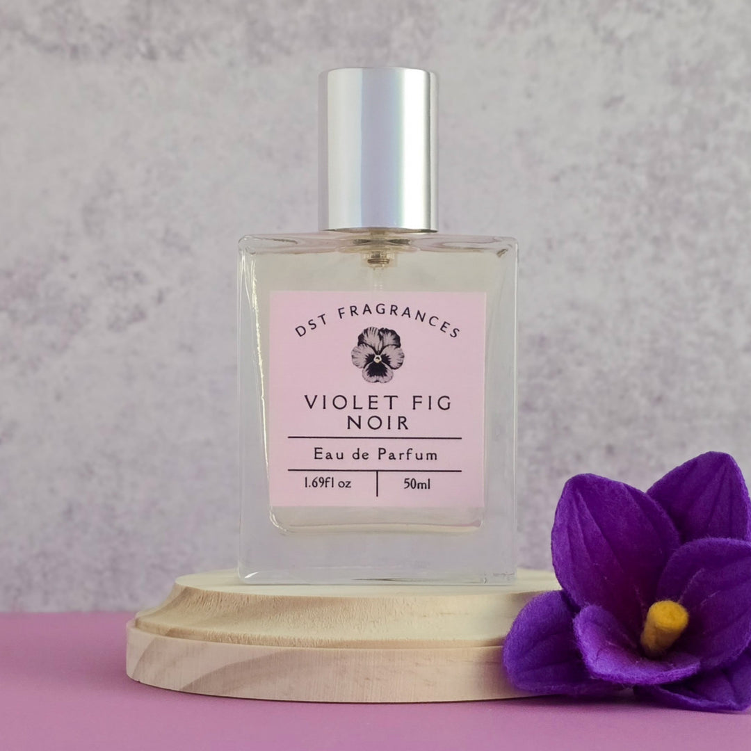 Perfume bottle labeled 'Violet Fig Noir' by DST Fragrances on a pink surface with a purple flower.