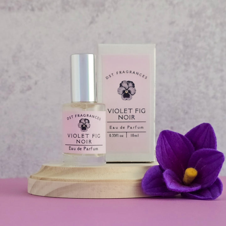 Violet Fig Noir perfume bottle and packaging on a pink surface with a purple flower.