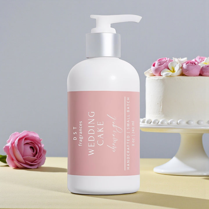 240ml bottle of Wedding Cake scented shower gel by DST Fragrances sitting on a pale-yellow surface with light grey background and a white wedding cake with Pink Roses.