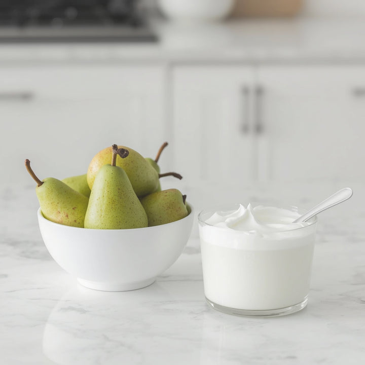 A glass bowl filled with green pears sits beside a small glass jar of white yogurt with a spoon on a light marble countertop in a bright kitchen.