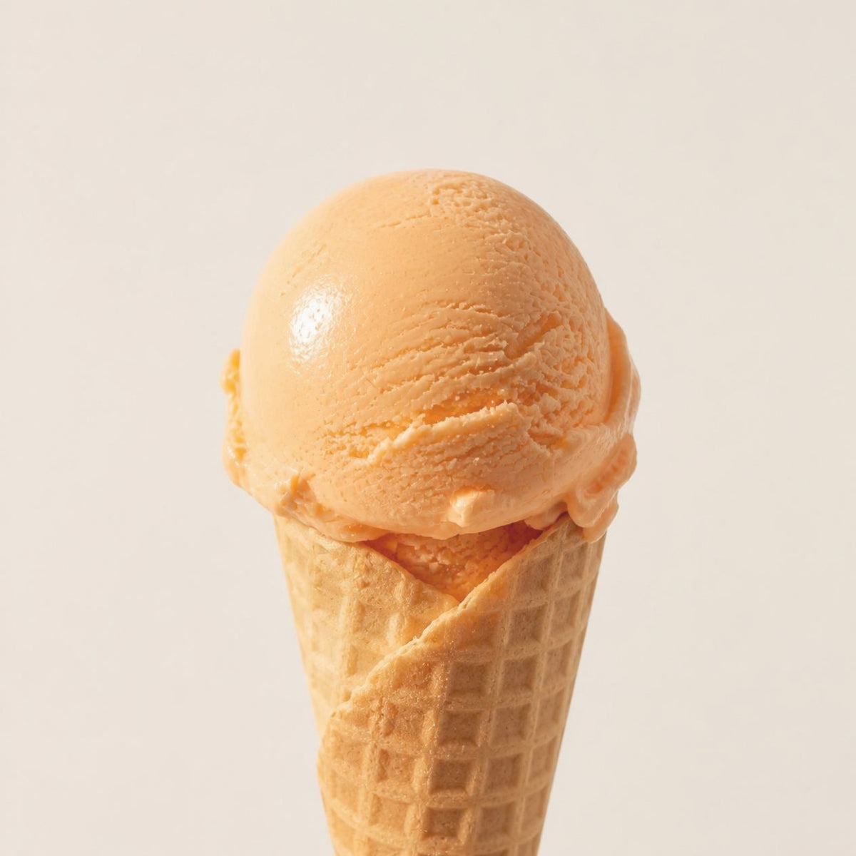 A scoop of light orange-cream ice cream in a classic waffle cone, photographed against a clean, solid, warm off-white background.