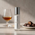 A clear glass perfume oil bottle labeled 'COGNAC & PRALINE' with a reflective silver cap, standing on a light surface. A blurred cognac glass and a plate of chocolates appear in the background, suggesting a fragrance note.