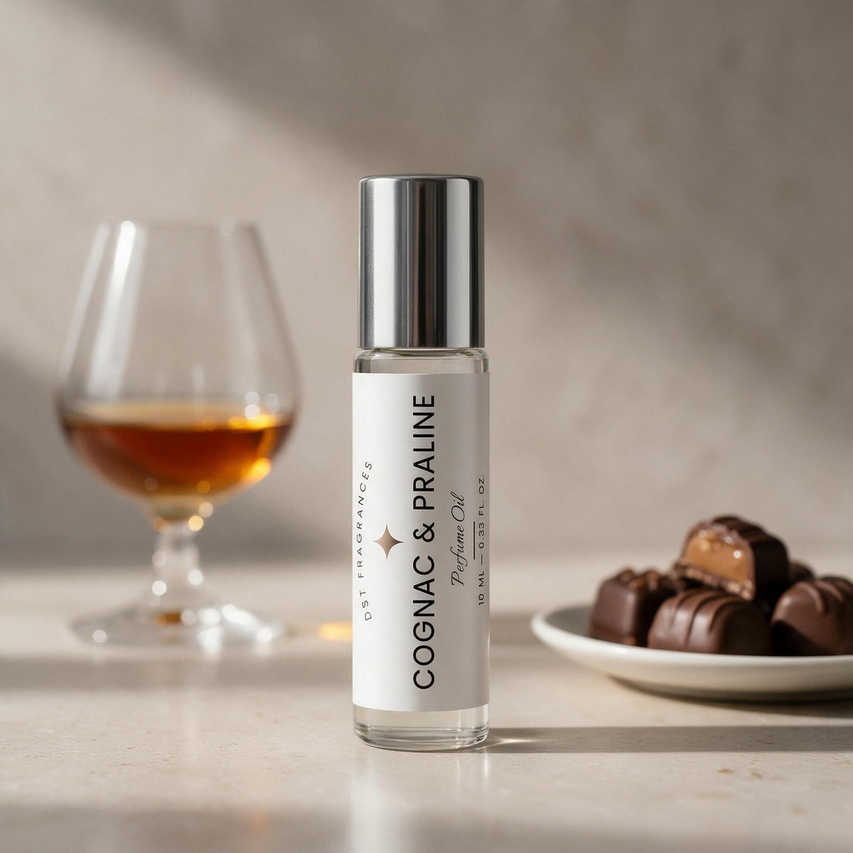 A clear glass perfume oil bottle labeled 'COGNAC & PRALINE' with a reflective silver cap, standing on a light surface. A blurred cognac glass and a plate of chocolates appear in the background, suggesting a fragrance note.